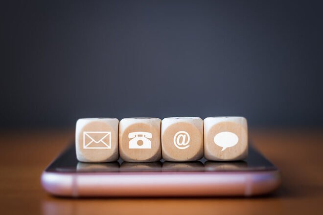 Drei hölzerne Würfelblöcke auf einem Smartphone. Auf jedem Würfel ist ein Symbol abgebildet: Telefon, Adresse, E-Mail, Sprechblase