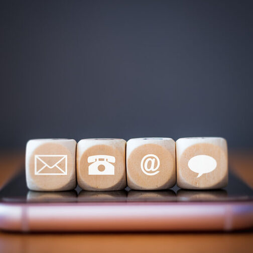 Three wooden dice blocks on a smartphone. Each cube has a symbol on it: Phone, address, e-mail, speech bubble.