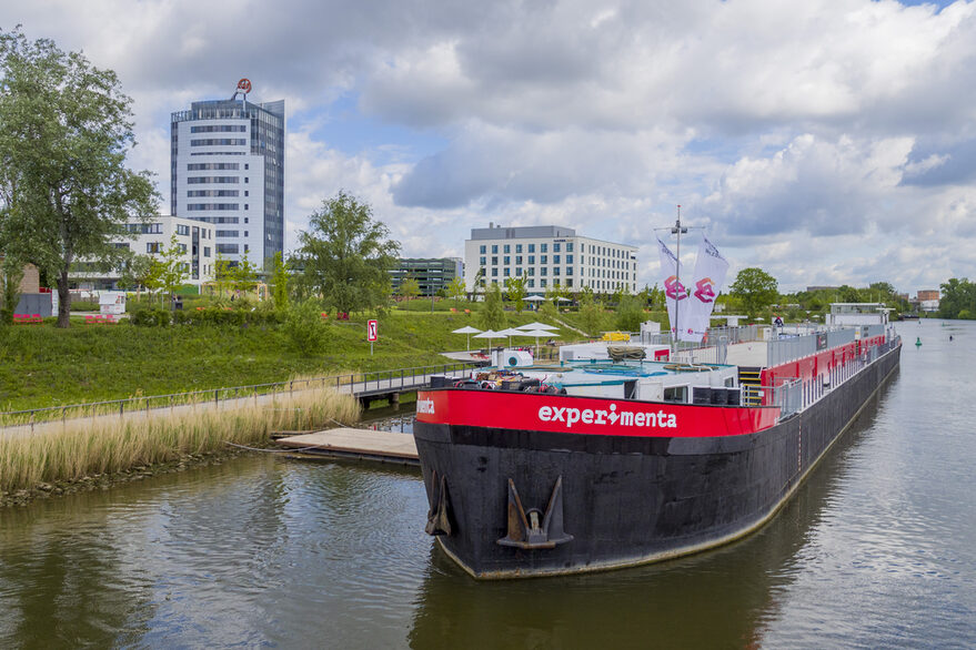 Das Schiff MS Experimenta im Neckarliegend, vor einem Hochhaus und begrüntem Ufer.